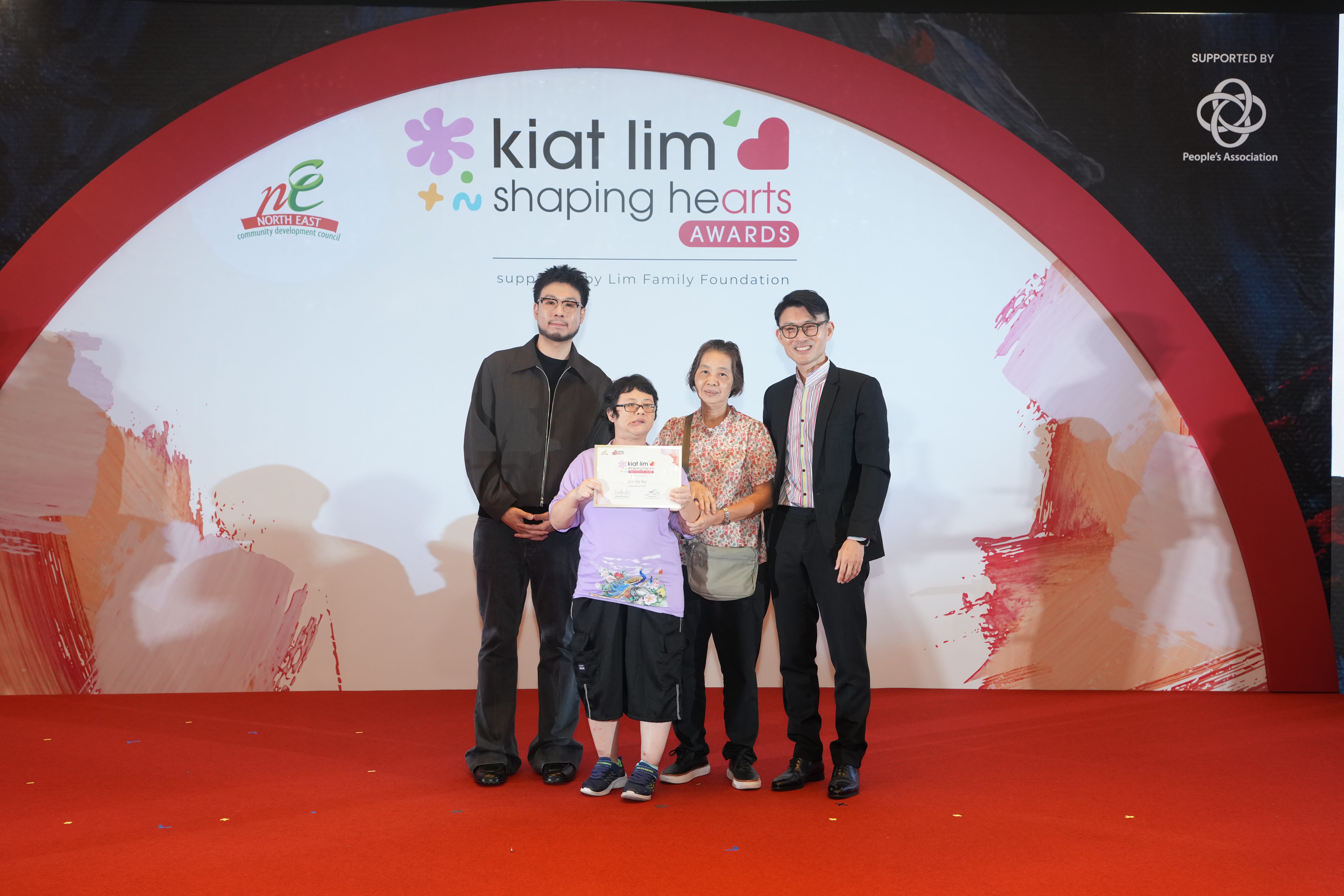 Award recipient standing on stage with the presenter, holding their certificate and posing for a group photo during the award ceremony, with a backdrop reading ‘Kiat Lim-Shaping Hearts Awards'.